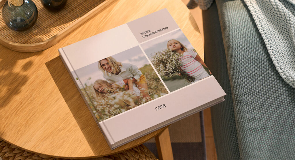 Auf einem Holztisch liegt ein CEWE FOTOBUCH mit dem Titel „UNSERE LIEBLINGSMOMENTE 2026“ und Fotos einer Mutter mit zwei Kindern.