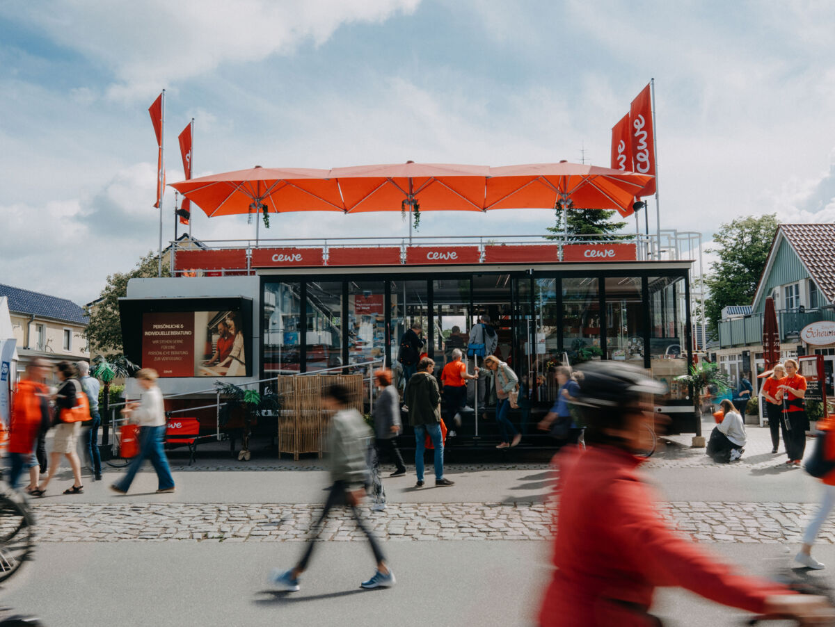 Der CEWE Truck steht bei Sonnenschein in der Einkaufsstraße. Im Vordergrund sind viele Passanten in Bewegung aufgenommen.