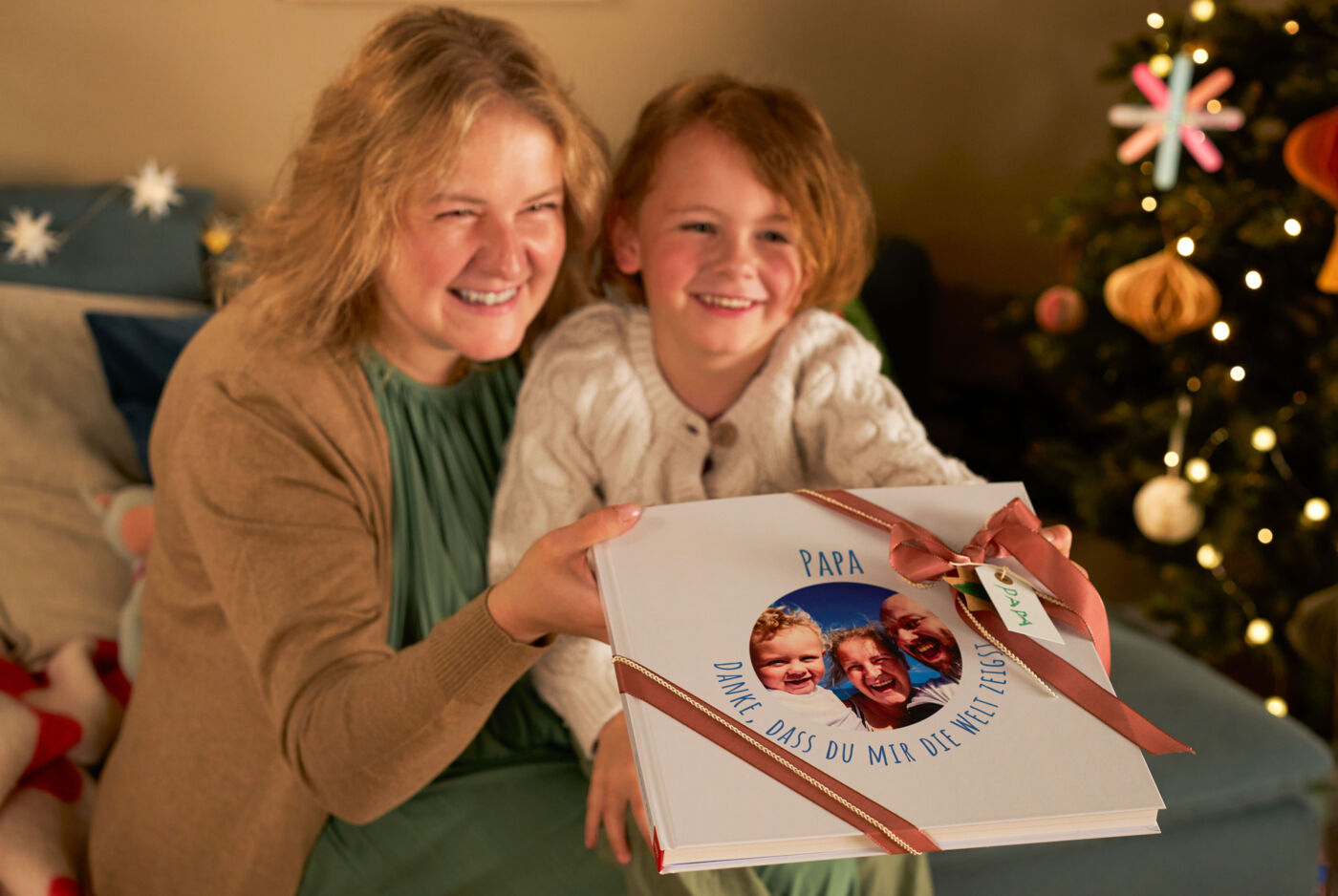 Eine Frau sitzt mit ihrem Kind vor einem Weihnachtsbaum. Beide lachen und halten ein CEWE FOTOBUCH in den Händen.