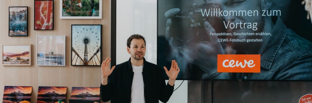 Aaron Moser hält während eines Vortrags die Hände erhoben und spricht vor einem Publikum, hinter ihm steht „Willkommen zum Vortrag"